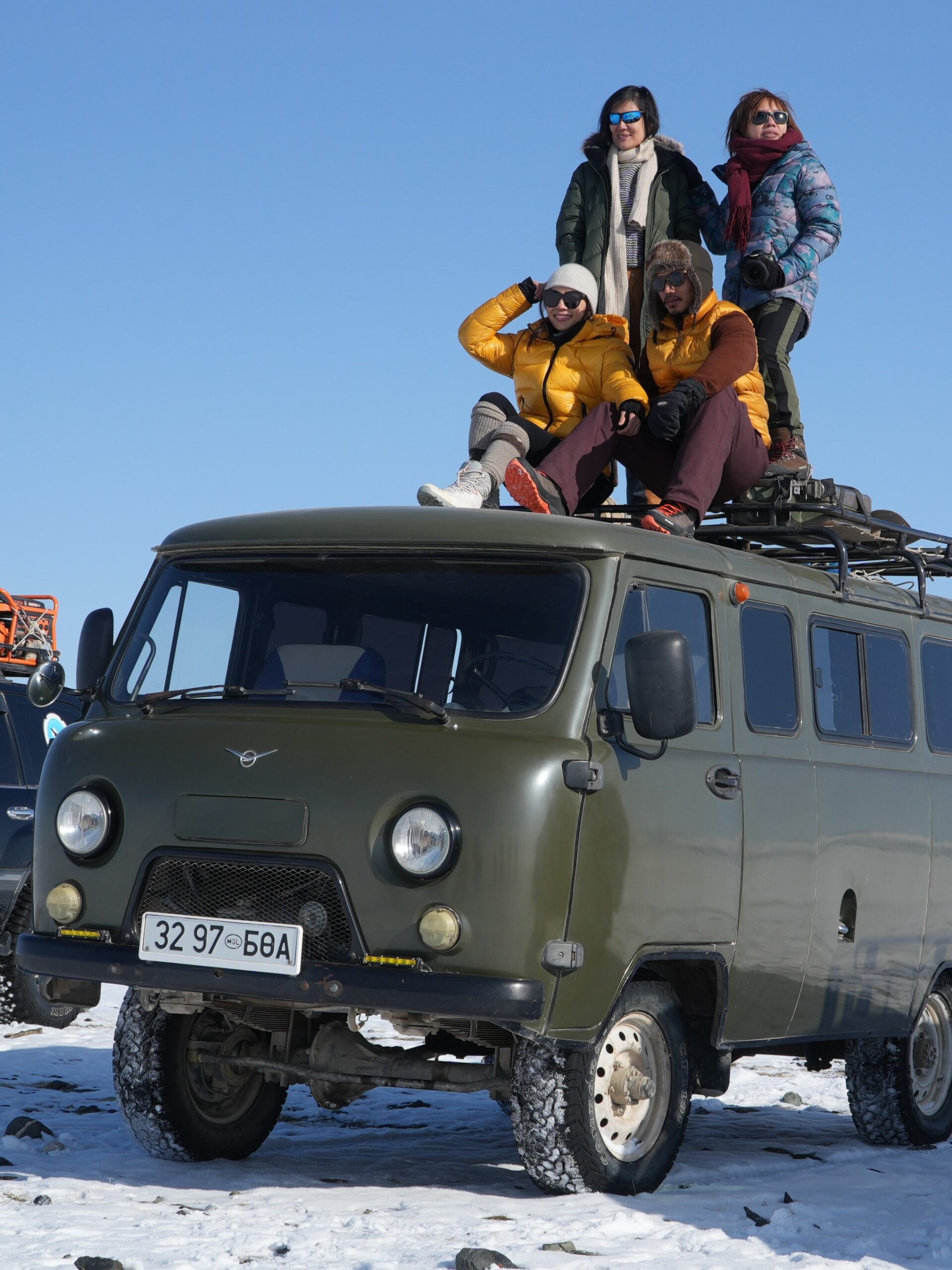 Four travellers sitting and standing on the roof of an olive-green UAZ van on snowy ground under a blue sky