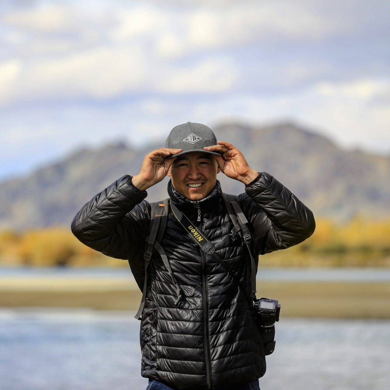 Yerlan Shakhabai smiling and adjusting his cap, with a camera on his shoulder, beside a river with autumn trees and mountains behind