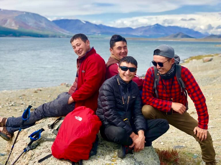 Four men from the Sayat Travel team laughing together on a boulder beside a mountain lake