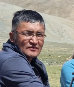 Portrait of Yerlan Kukyebai in glasses and a navy jacket, with bare hills behind