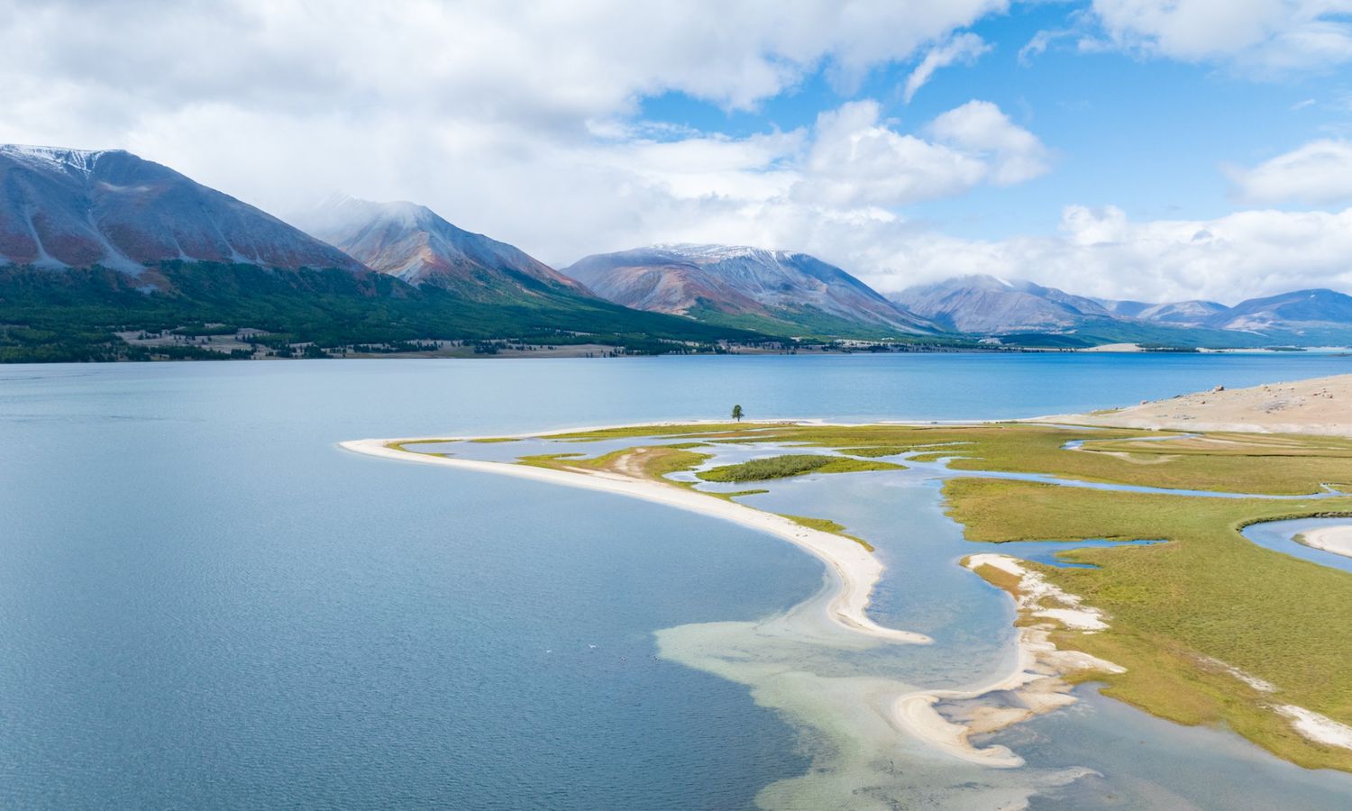 Aerial view of a blue mountain lake with a curving white sand spit and green marsh, below red and green mountains