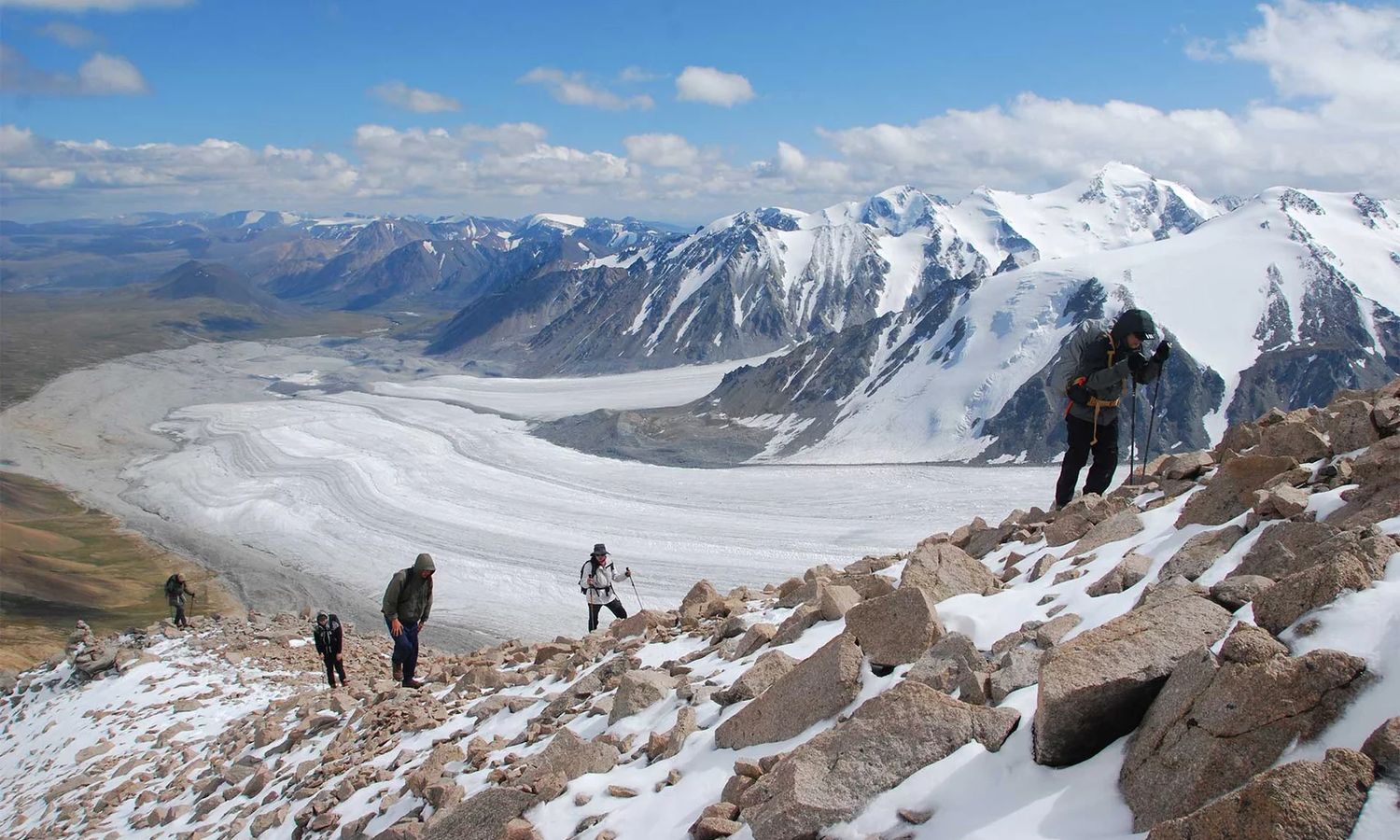 A small group trekking up a snowy rock ridge above a wide glacier and snow peaks in Altai Tavan Bogd National Park
