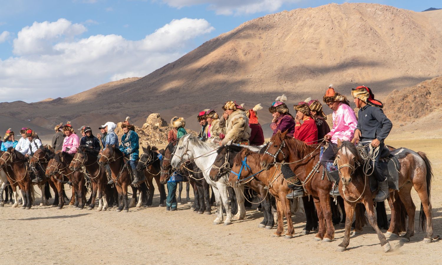 A long line of eagle hunters on horseback in traditional dress at an eagle festival, with dry mountains behind