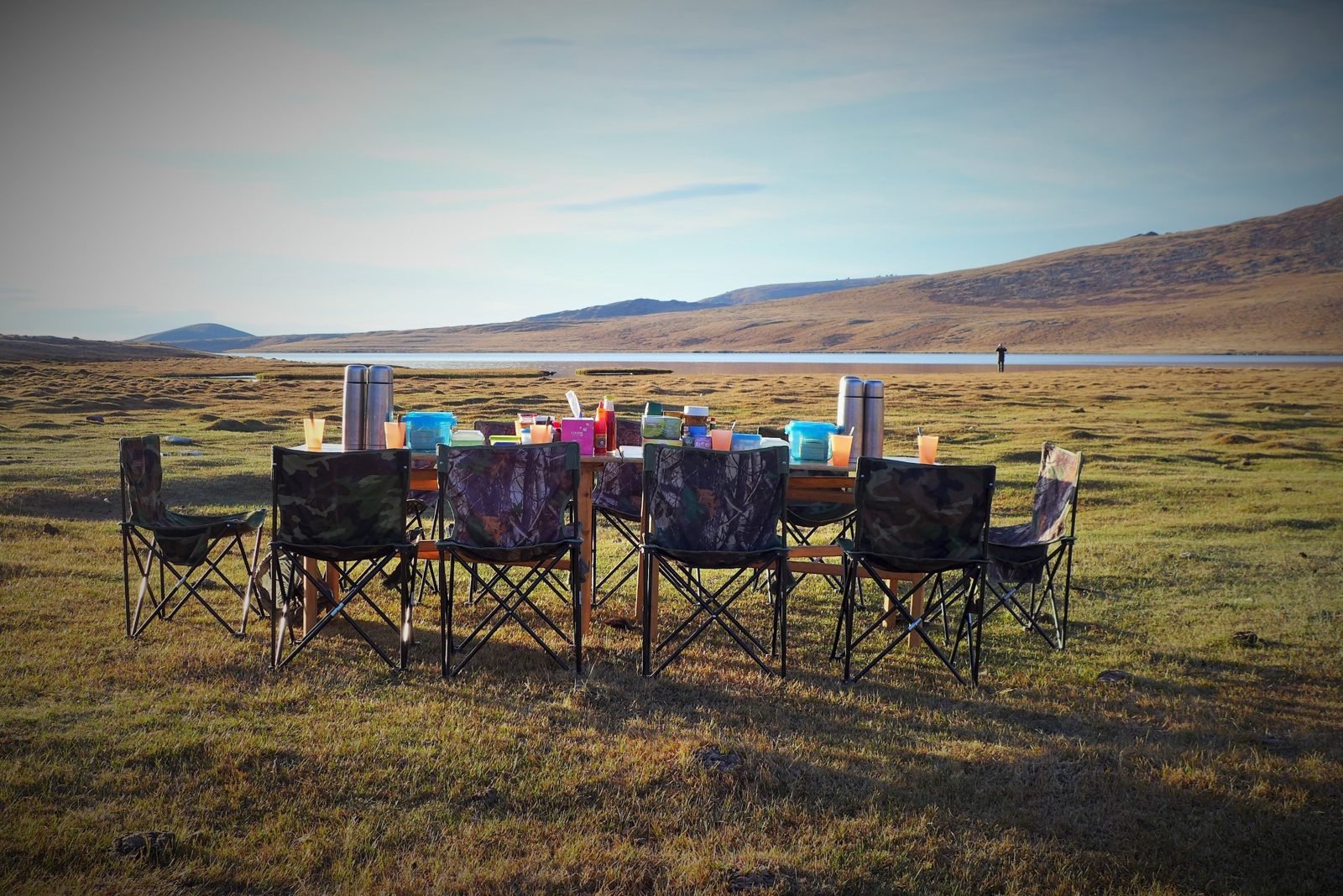 A long camp dining table with thermos flasks, cups and camouflage folding chairs set out on grassland beside a lake at sunrise