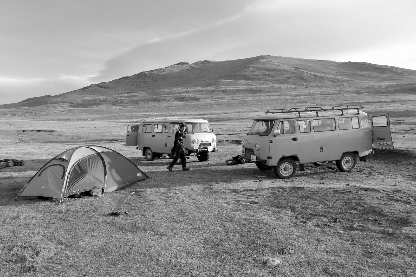 Black-and-white photo of a dome tent and two UAZ vans with open doors on open steppe below a bare hill
