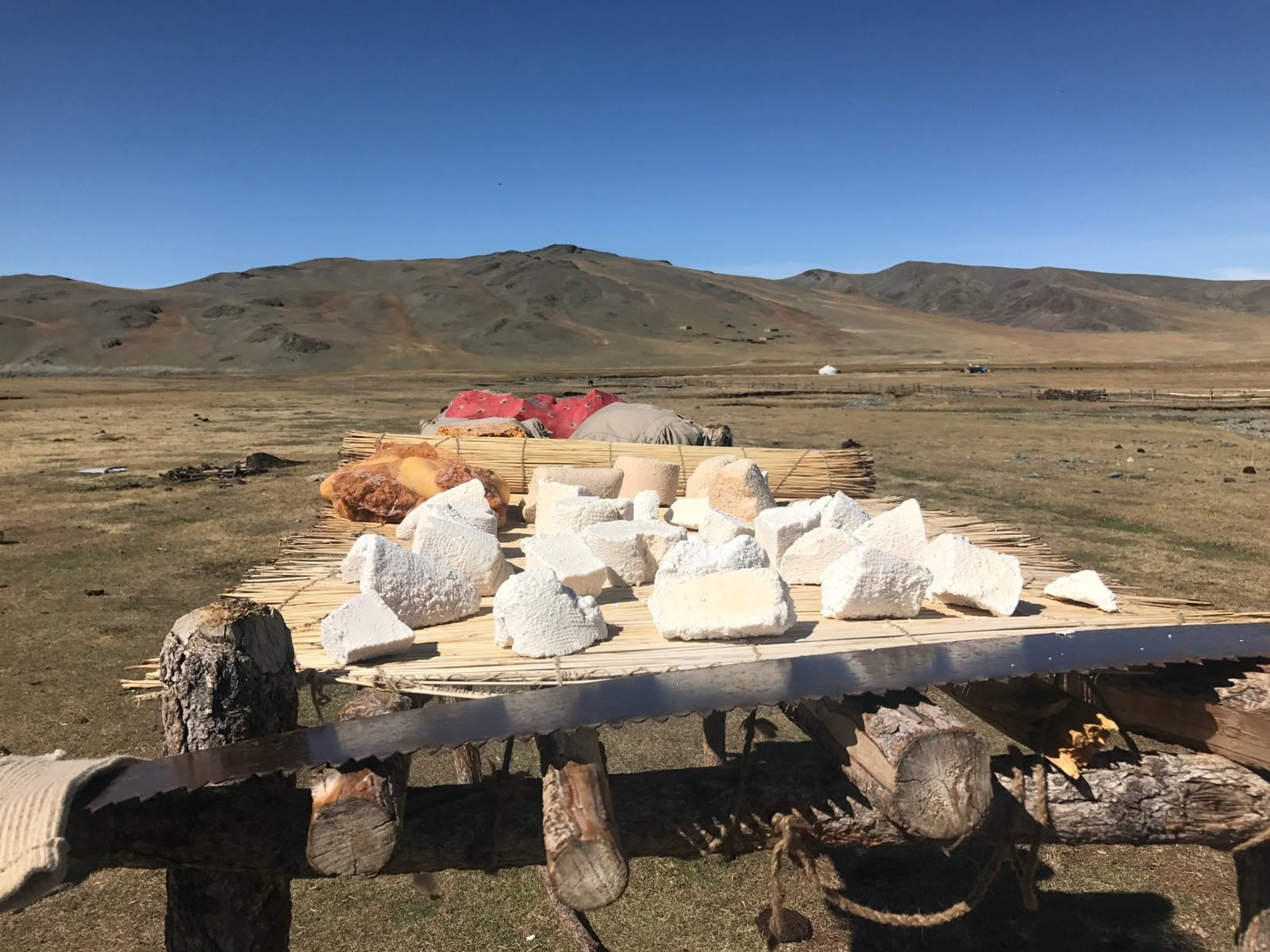 Pieces of white aaruul, dried curd, laid out on a reed mat on a wooden rack under a blue sky, with brown hills behind