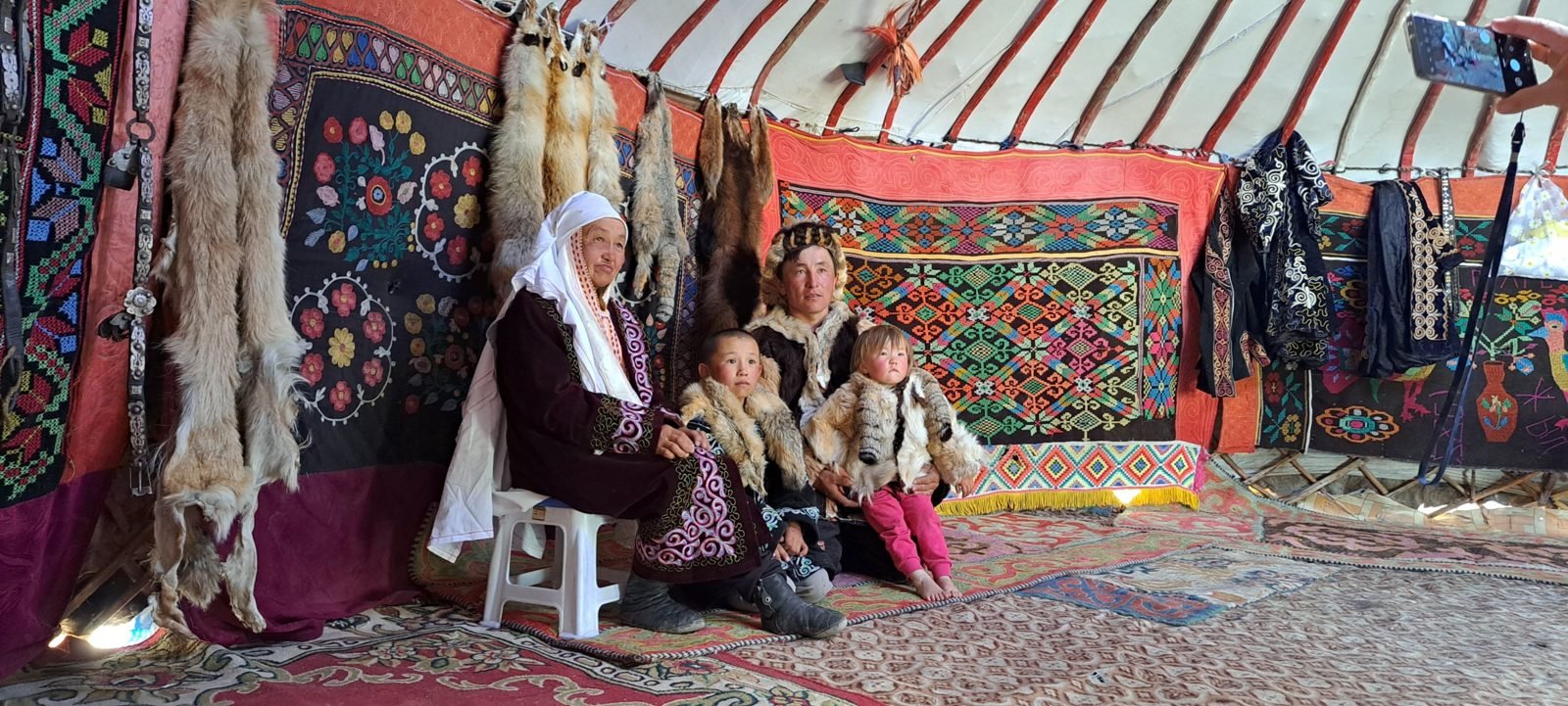 A Kazakh eagle hunter in a fox-fur hat with his mother and two small children in fur coats, sitting in front of embroidered wall hangings and fox pelts in their ger