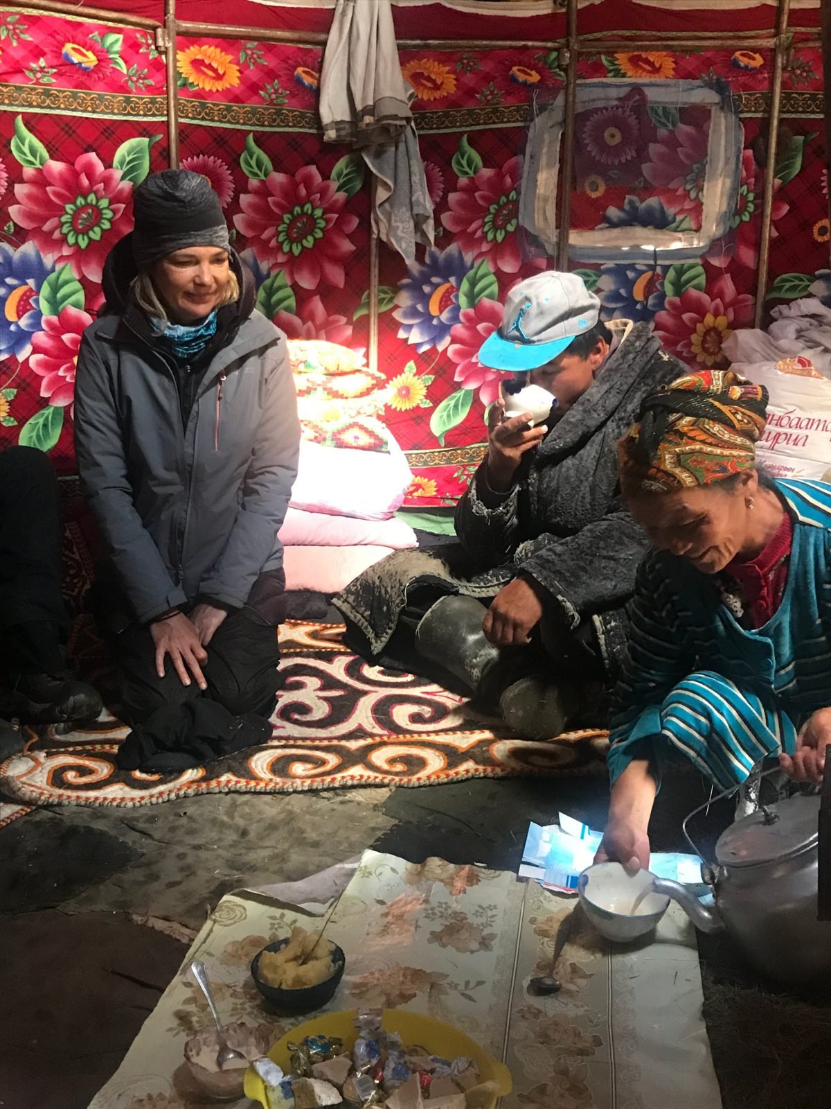 A smiling guest kneels on a felt rug in a ger hung with floral fabric while a herder drinks tea and an older woman pours milk tea from a kettle
