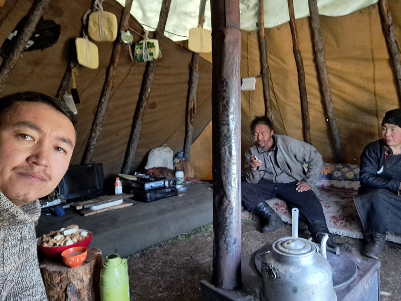 A man takes a selfie inside a herders' canvas tent while an older herding couple sit by the stove and kettle