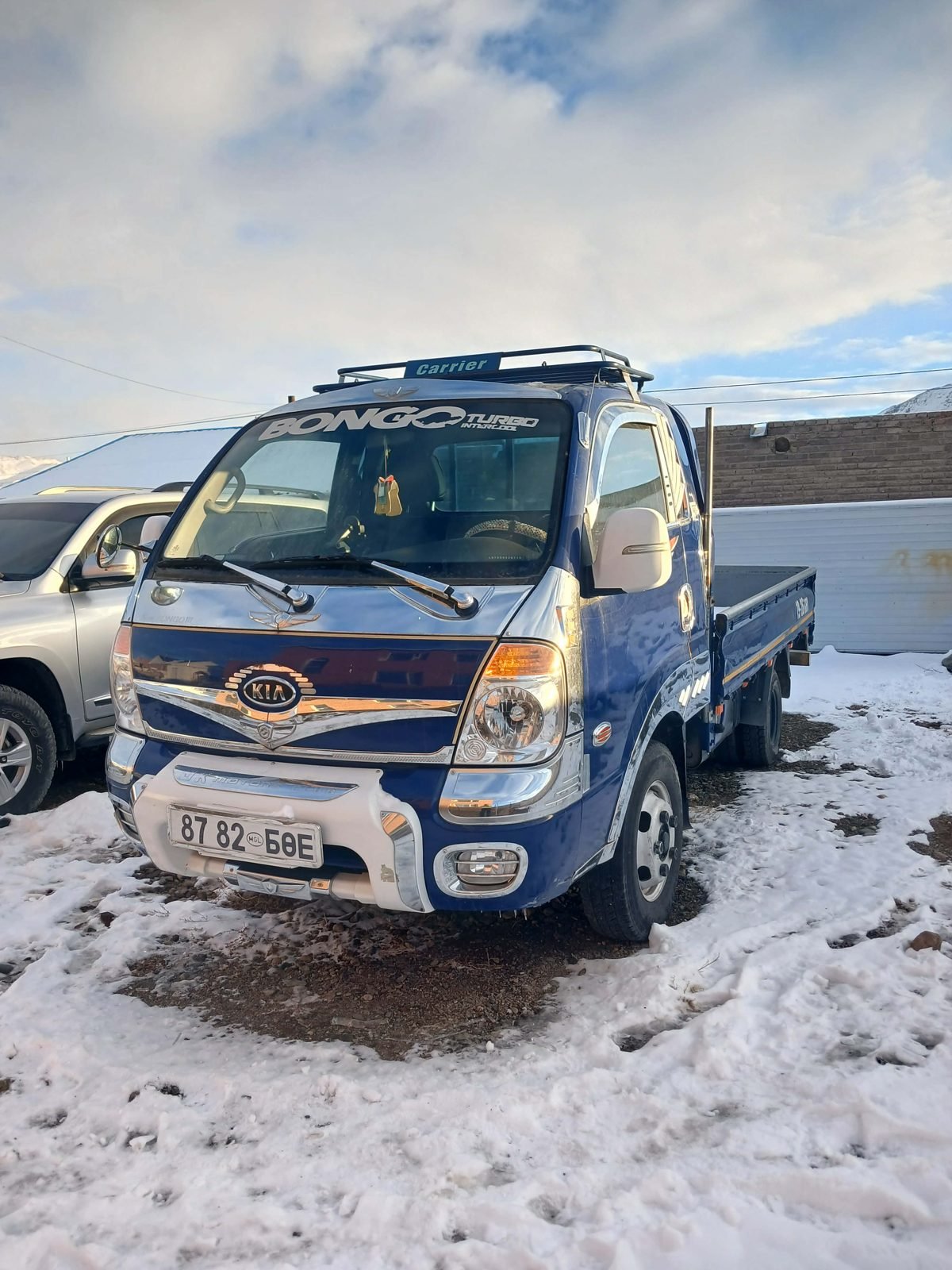 A blue Kia Bongo minitruck with a roof rack and an open cargo bed, parked on snow