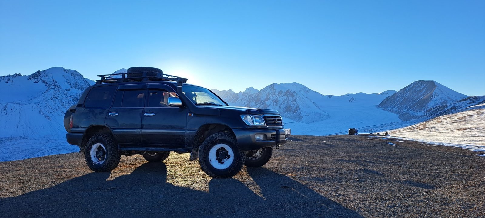 Our black Toyota Land Cruiser 105 with a roof rack and snorkel parked on a high gravel pass in front of a glacier and snow-covered peaks