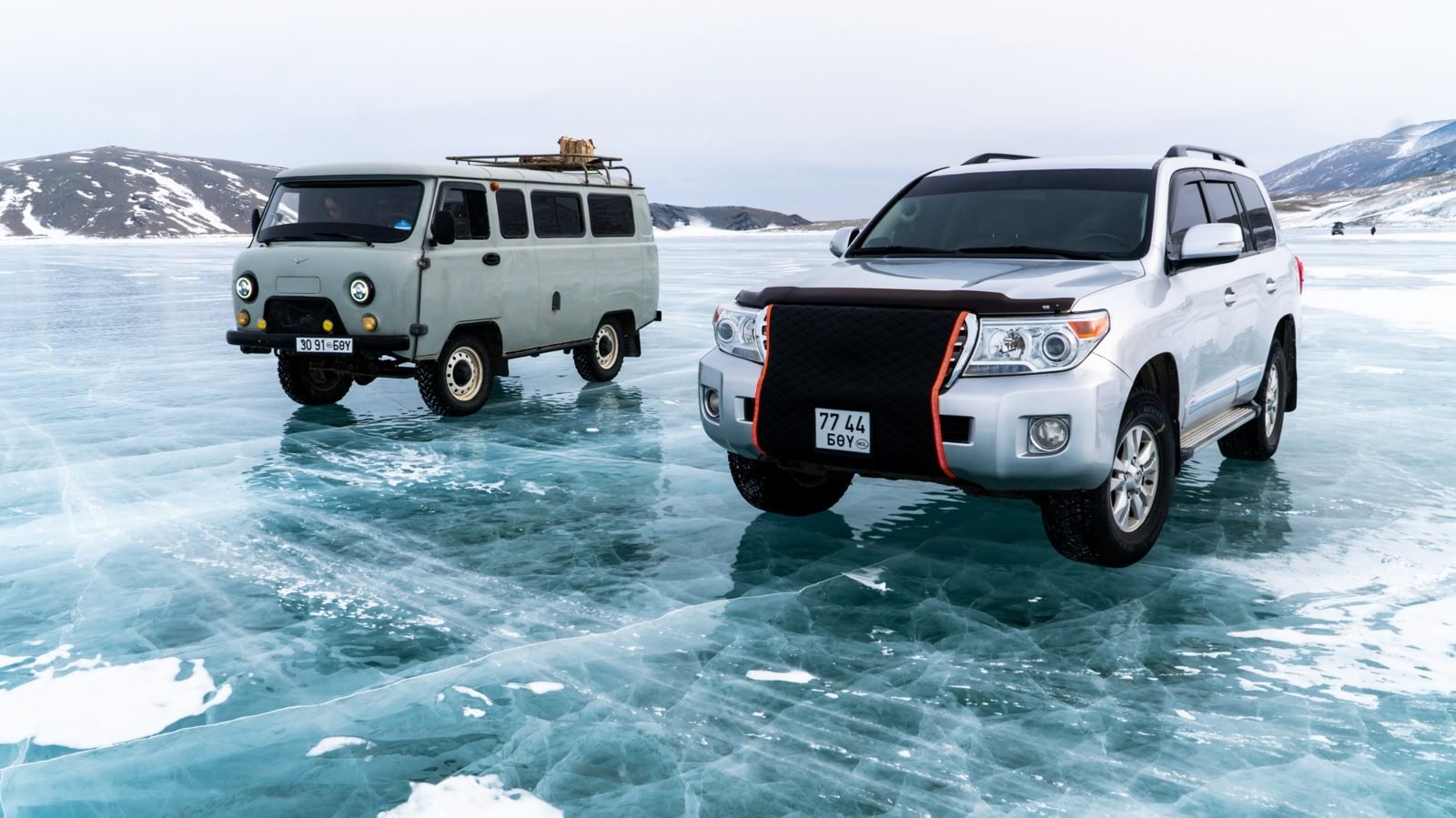 Our silver Toyota Land Cruiser 200 with a winter radiator cover parked on clear blue ice on a frozen lake, beside a UAZ van