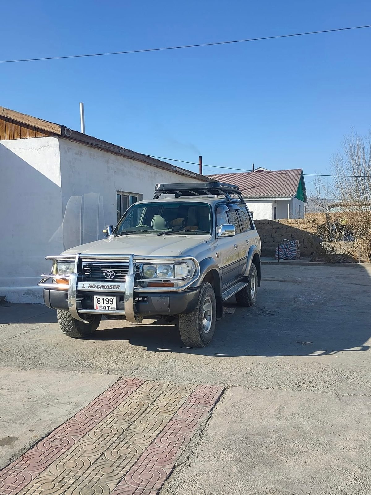 A silver Toyota Land Cruiser 80 with a roof rack and chrome bull bar parked in a sunny yard beside a white building