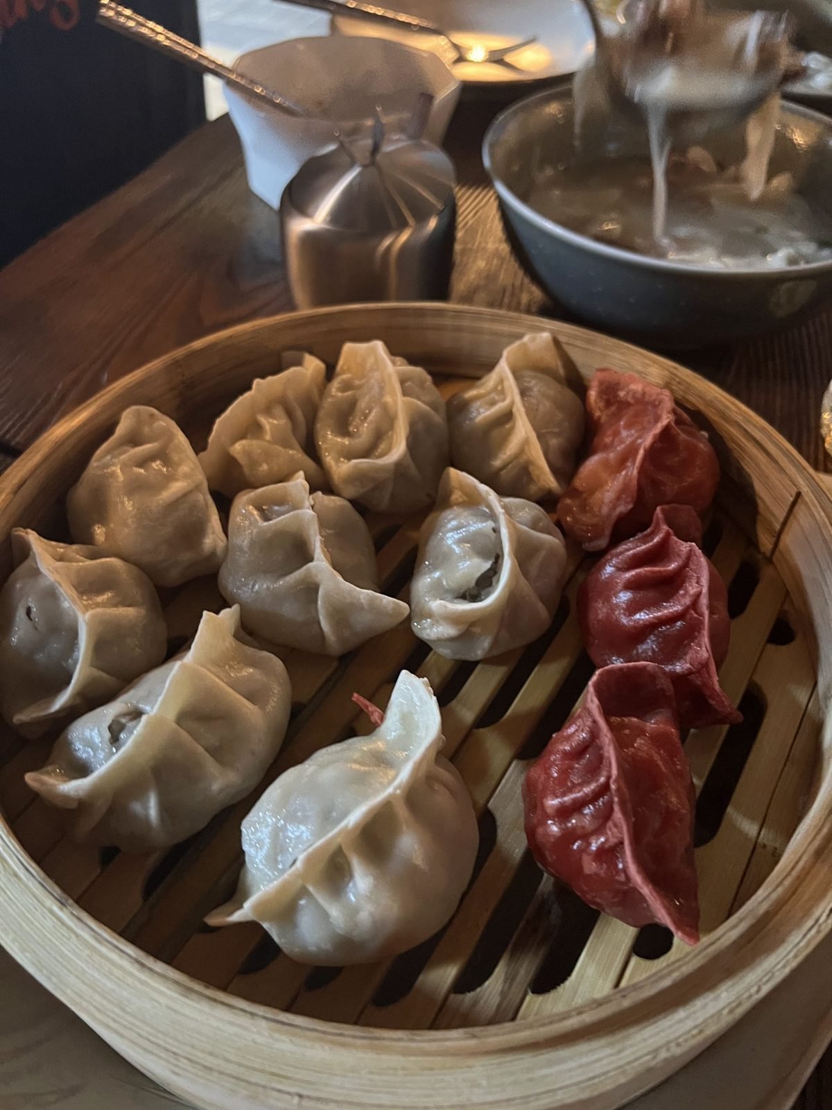 A bamboo steamer of buuz dumplings, some made with red dough, beside a pot of soup