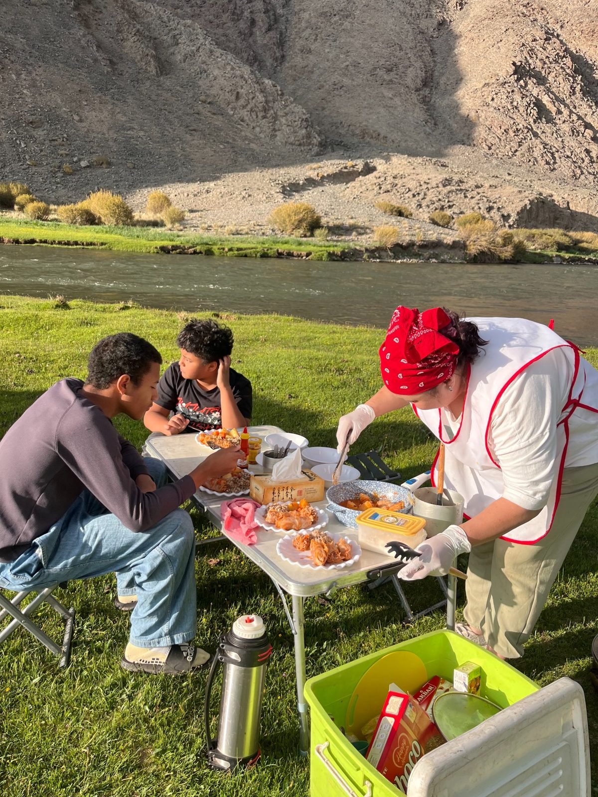 Our cook in a red bandana and apron serving stuffed cabbage rolls and potatoes to young guests at a folding table beside a river