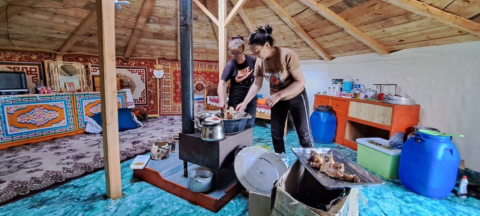 Two women preparing meat in a large pan on the stove of a wooden-roofed ger