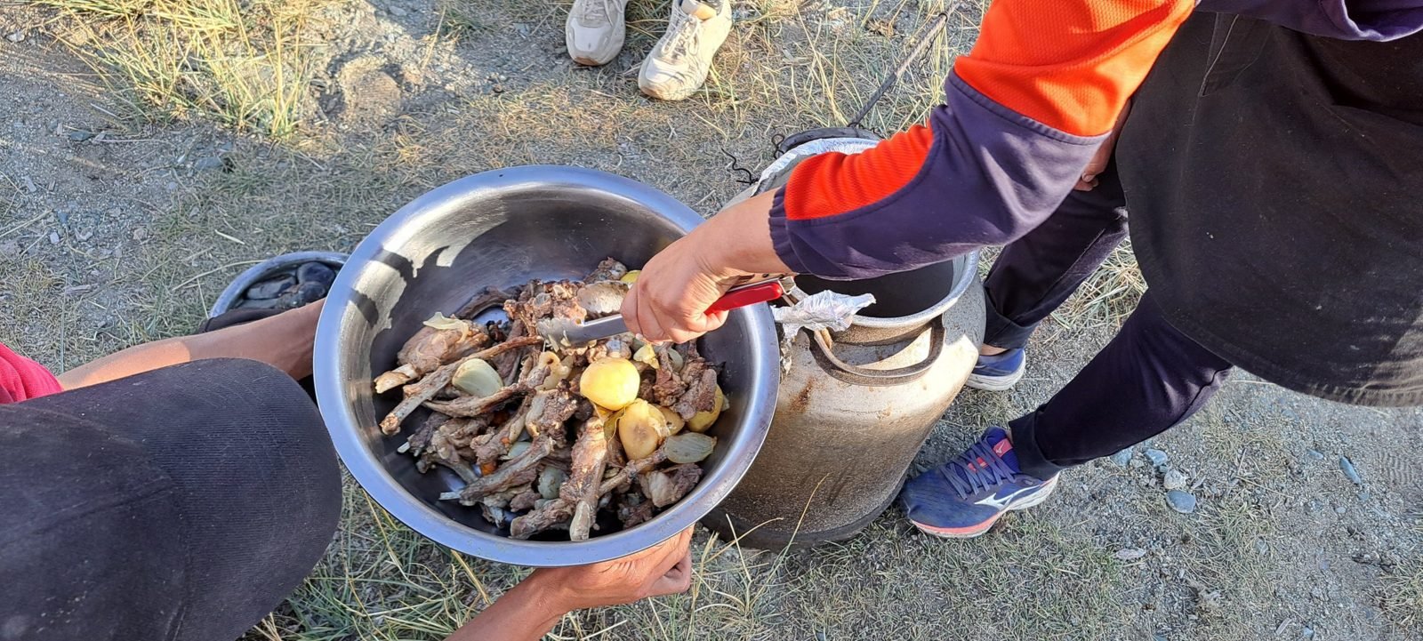Tongs lifting pieces of mutton, potatoes and onion out of a milk can into a steel bowl