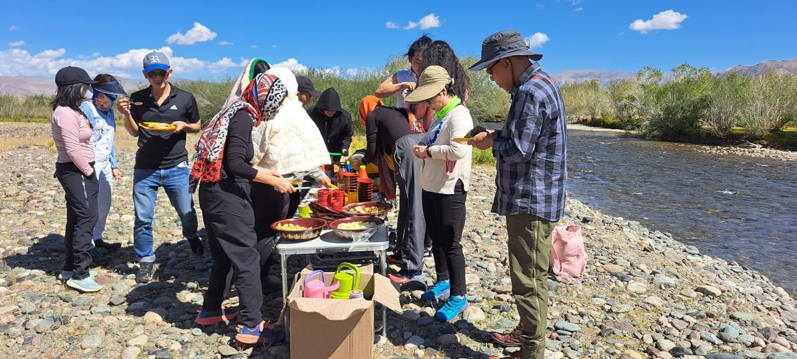 A group of travellers in sun hats serving themselves from bowls on a folding table on the stony bank of a fast river