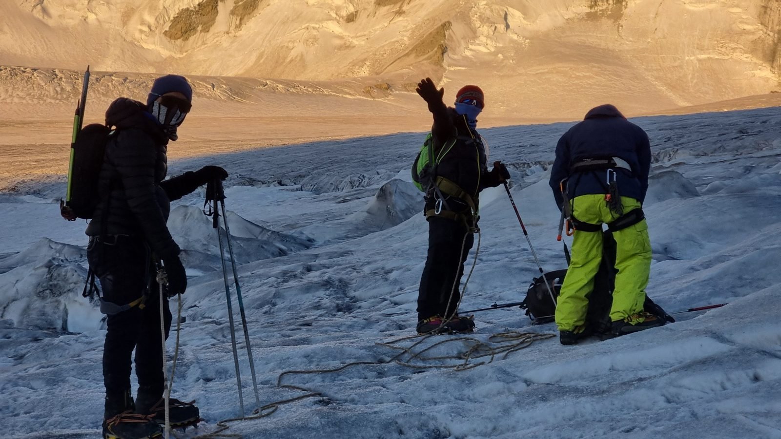 A roped team in harnesses and crampons on a glacier at sunrise, one climber raising an arm