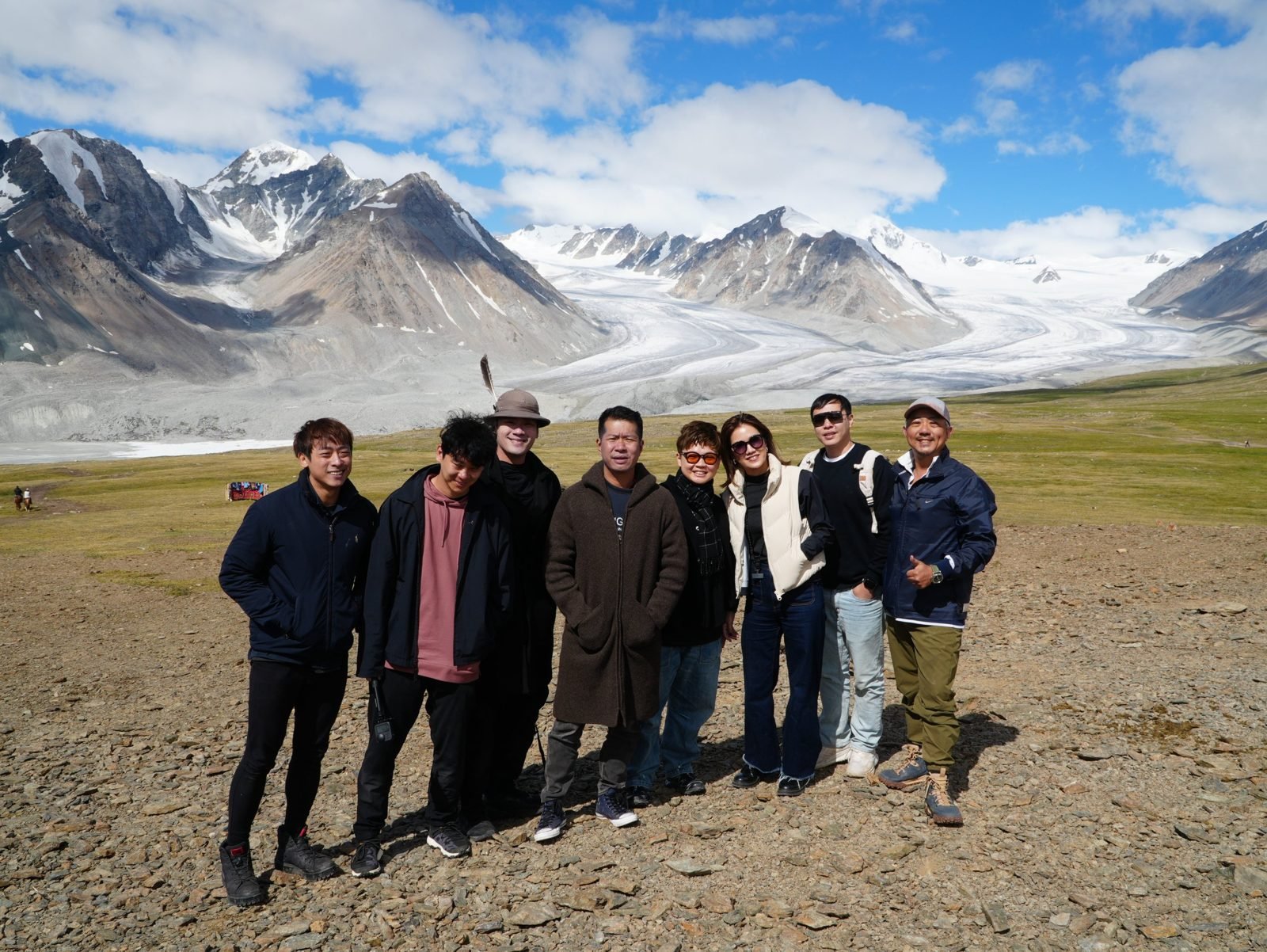 A group of eight travellers posing on stony ground in front of a huge glacier and snowy peaks in Altai Tavan Bogd