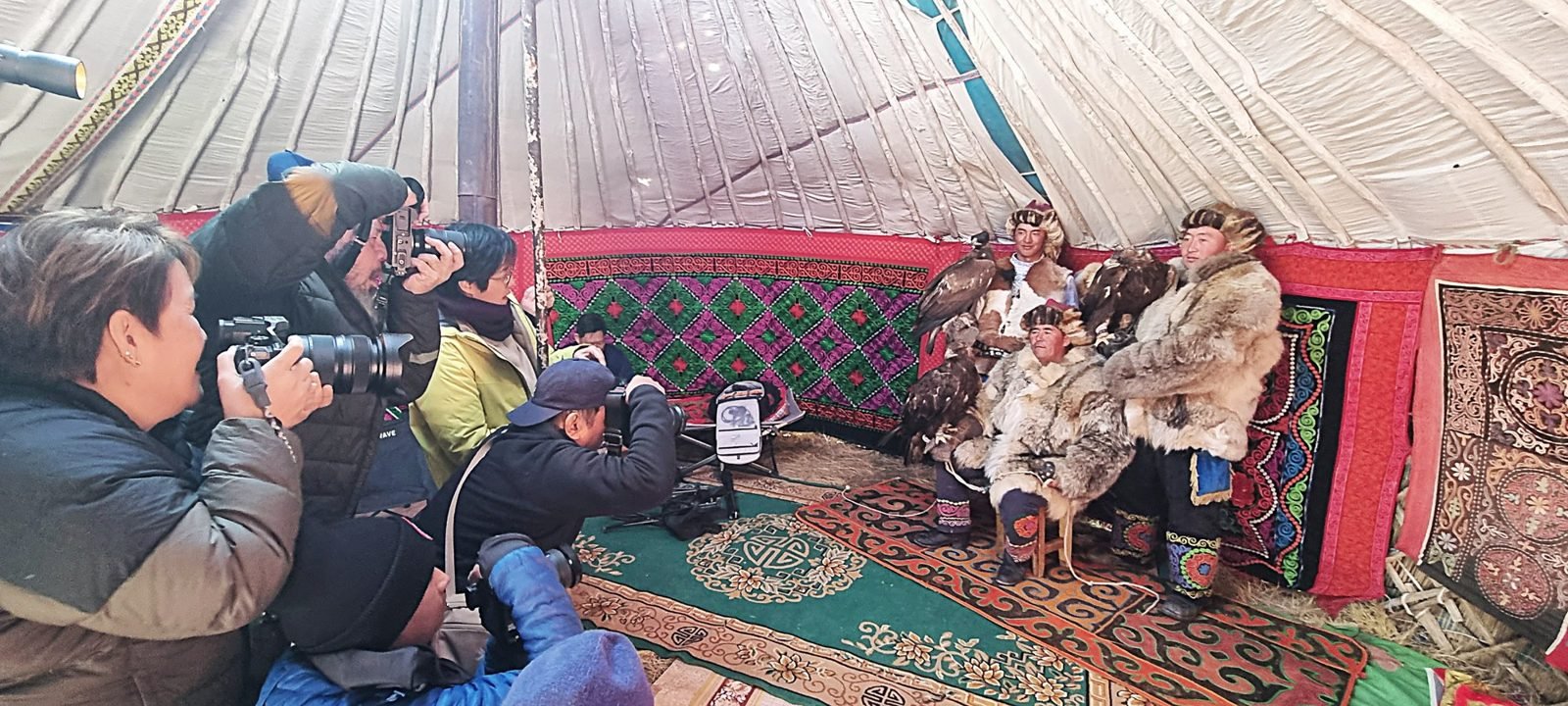 Photographers with cameras and long lenses kneeling inside a ger to photograph two Kazakh eagle hunters in fur coats with their golden eagles