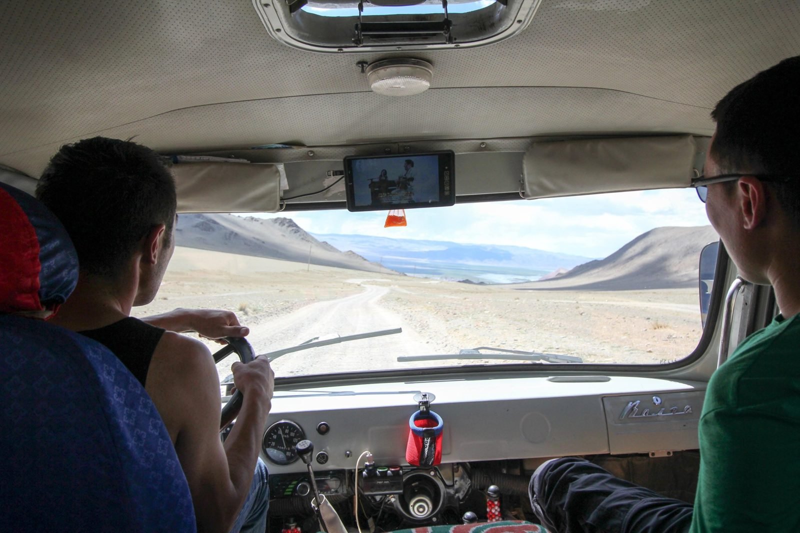View from inside a van over the driver's shoulder down a gravel track into a wide valley with a lake and bare mountains