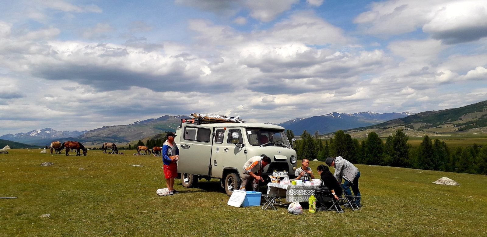 A grey UAZ van parked in a green Altai valley while the crew sets out a picnic lunch on a folding table, with horses grazing and snowy mountains behind