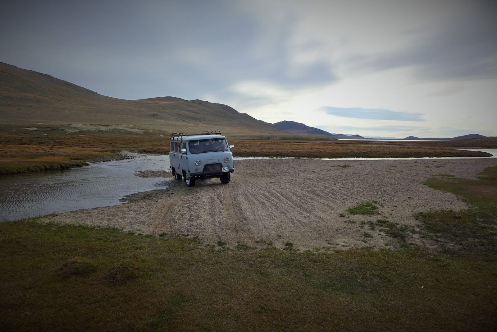 A pale blue UAZ van driving out of a shallow river onto a gravel bank, with brown hills and a lake behind