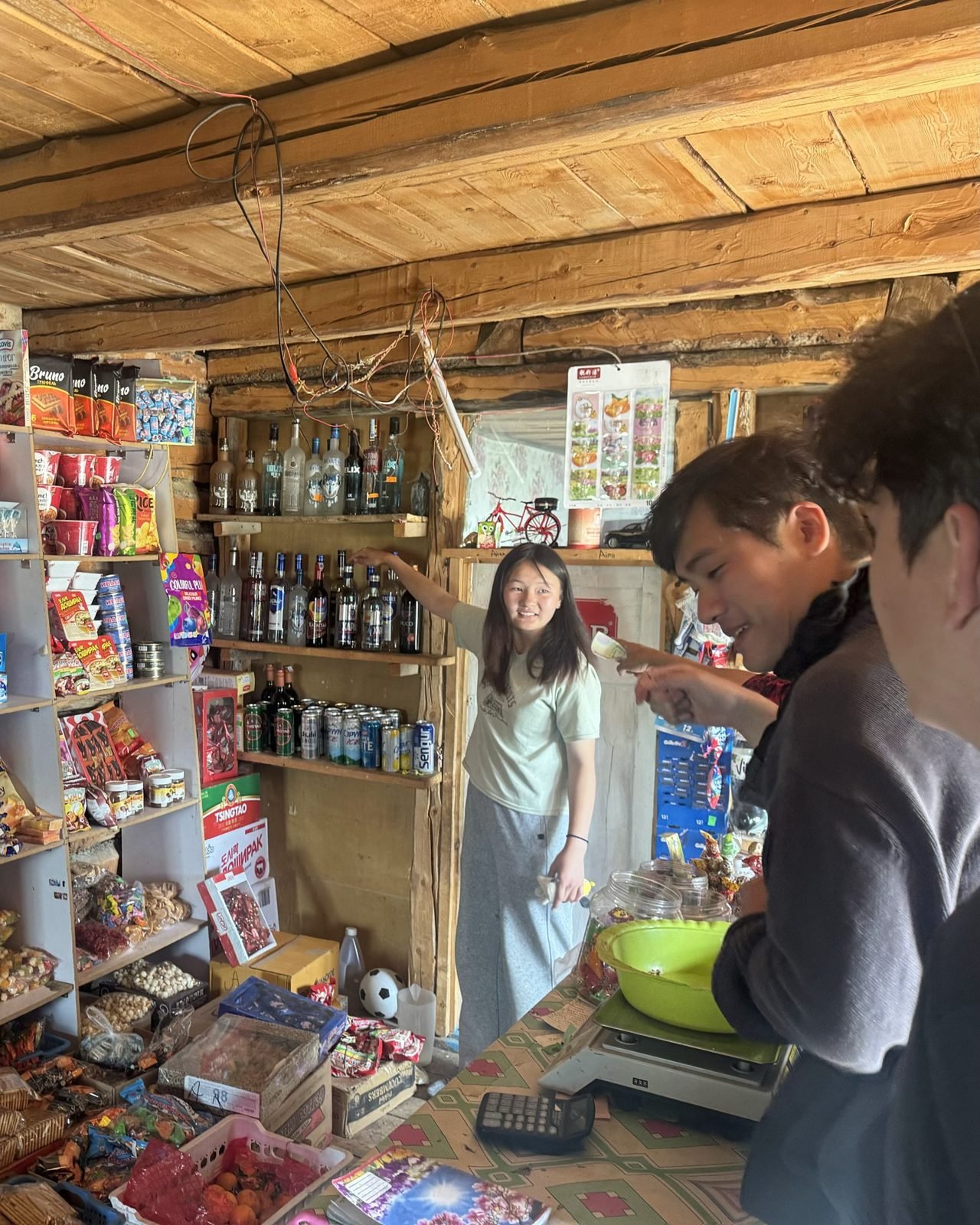 Travellers laughing at the counter of a small wooden village shop stocked with snacks and drinks