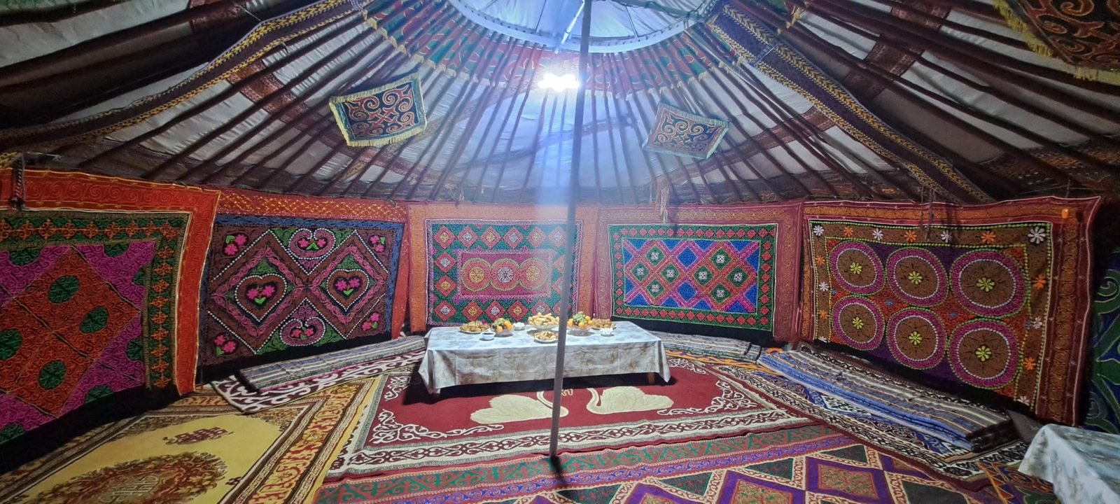 Inside a Kazakh ger hung with brightly embroidered felt wall hangings, with patterned rugs and a low table of food
