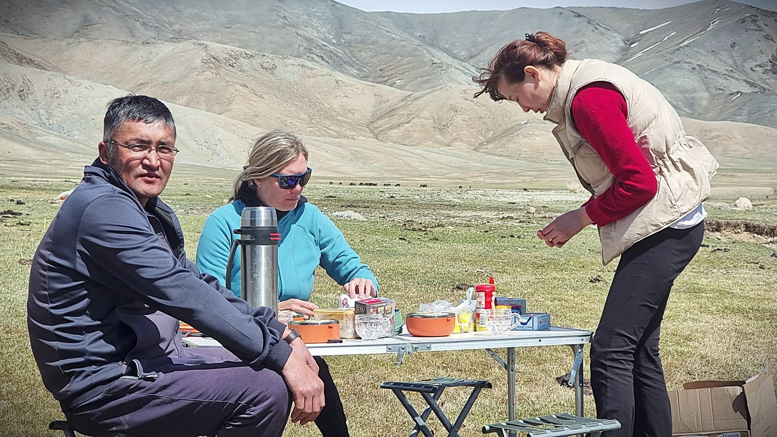 Our cook in a red jumper and fleece vest preparing lunch at a folding table on the steppe, where a team member and a guest sit with a thermos, with mountains behind