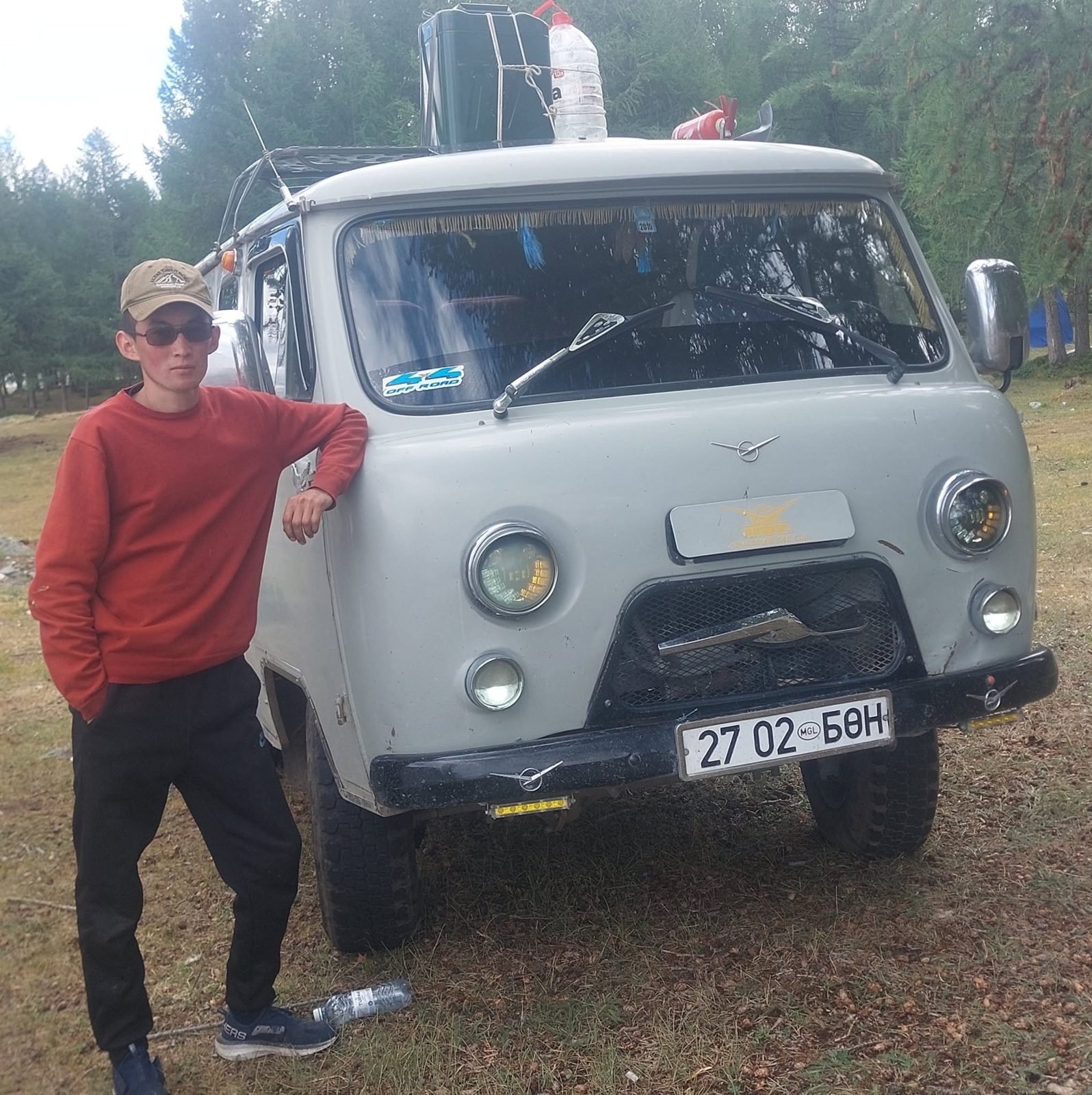 A driver in a rust-red sweater and cap leaning on the front of a grey UAZ van with fuel cans on the roof, among larch trees