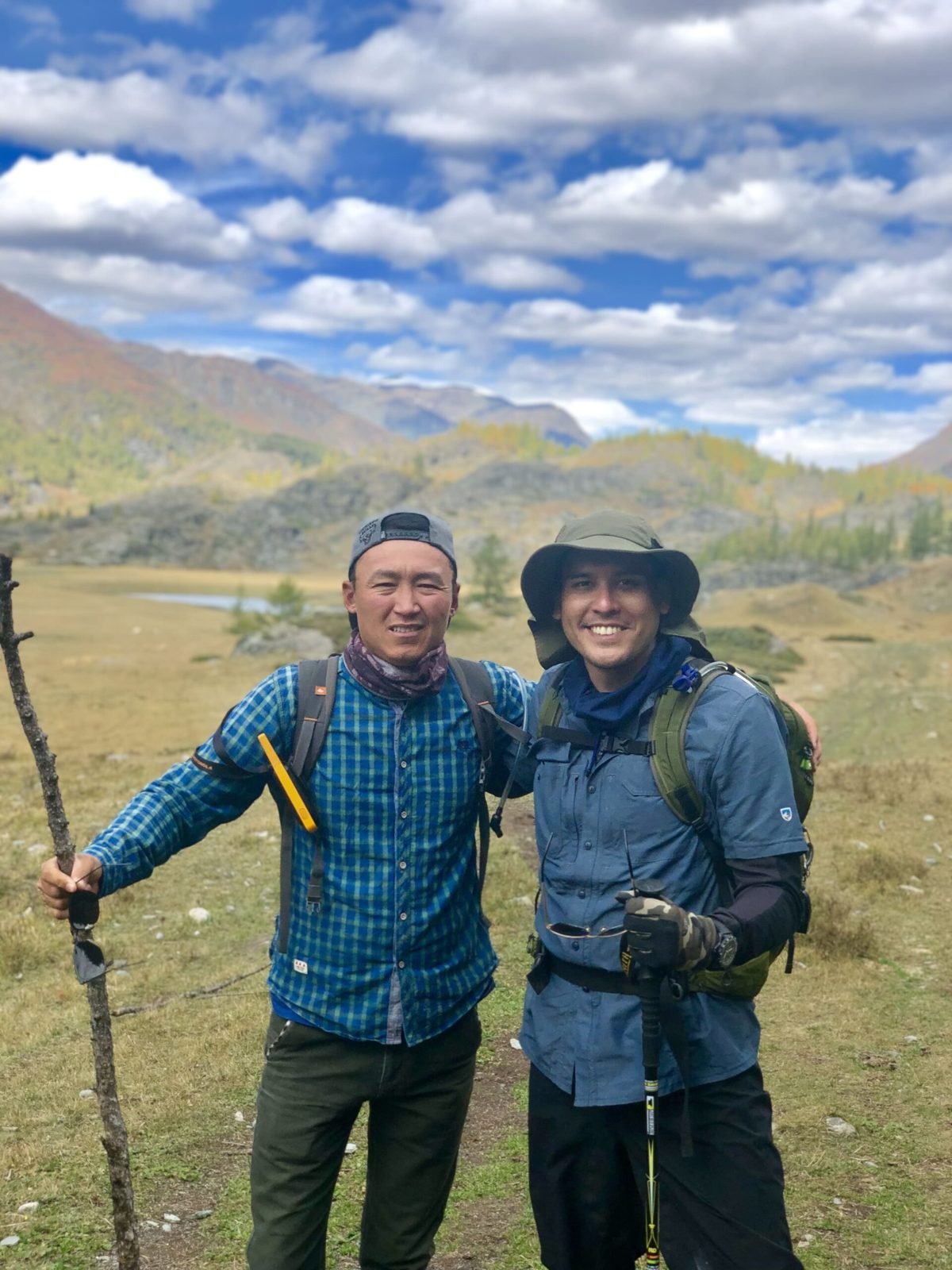 A guide in a blue checked shirt holding a wooden walking stick, arm round a smiling trekker in a sun hat, in an autumn mountain valley