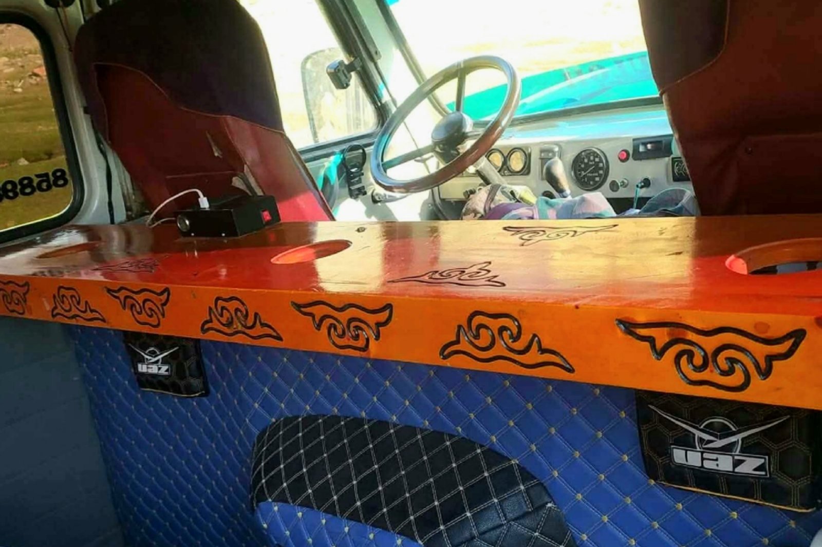 Inside our UAZ van: a hand-carved wooden console painted orange with Kazakh patterns, quilted blue panels with UAZ badges, and the driver's seat and wheel beyond